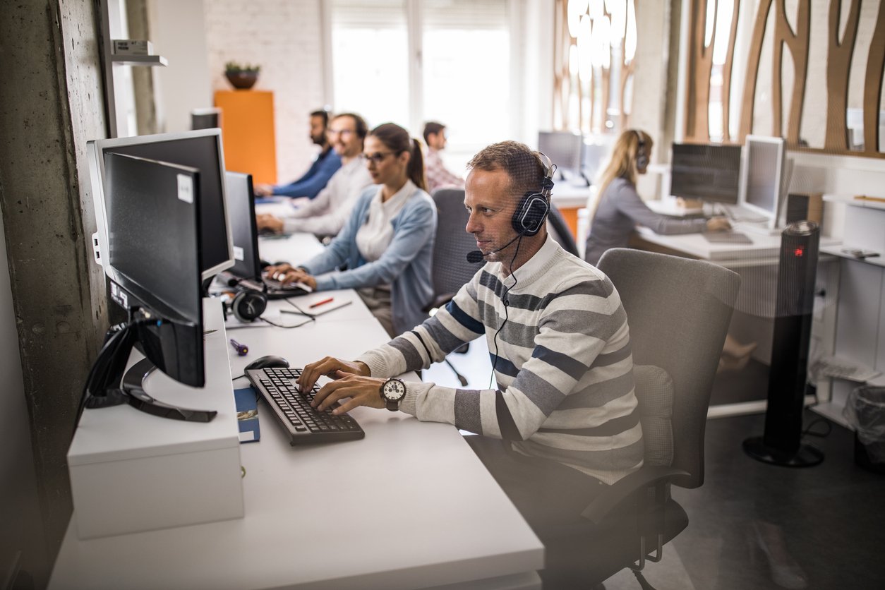 Young man working on desktop PC at call centre with his colleagues. The view is through glass.