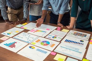 Financial and business documents on table with multiethnic hands working on it. Latin business manager with colleagues working on new startup project. Closeup business man and businesswoman hands understanding pie and bar graphs during meeting.