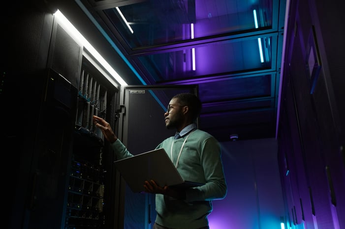 Low angle portrait of young African American data engineer working with supercomputer in server room lit by blue light and holding laptop, copy space