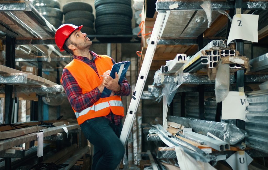 Worker doing audit in a glass cutting workshop. He started audit by doing inventory in the warehouse. In the background we can see batters needed in glass cutting business.